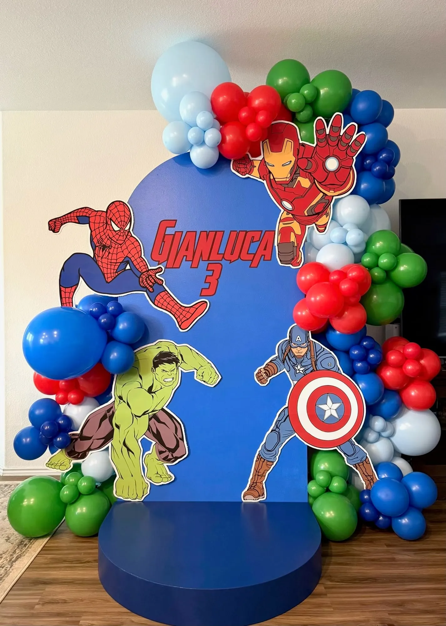 Superhero themed third birthday with a blue arch backdrop, hero cutouts, round stage and red, blue and green balloon garland
