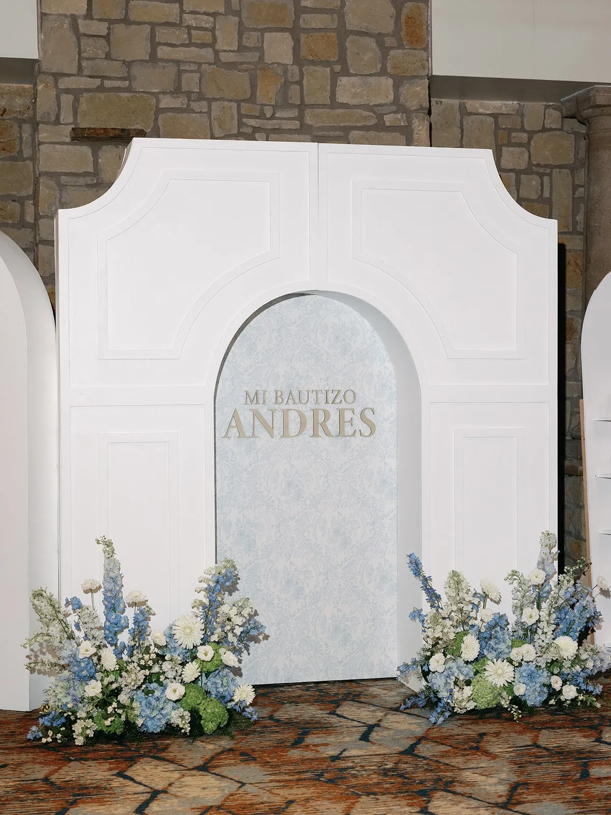 Baptism backdrop with a white paneled arch, blue toile insert and blue and white floral arrangements