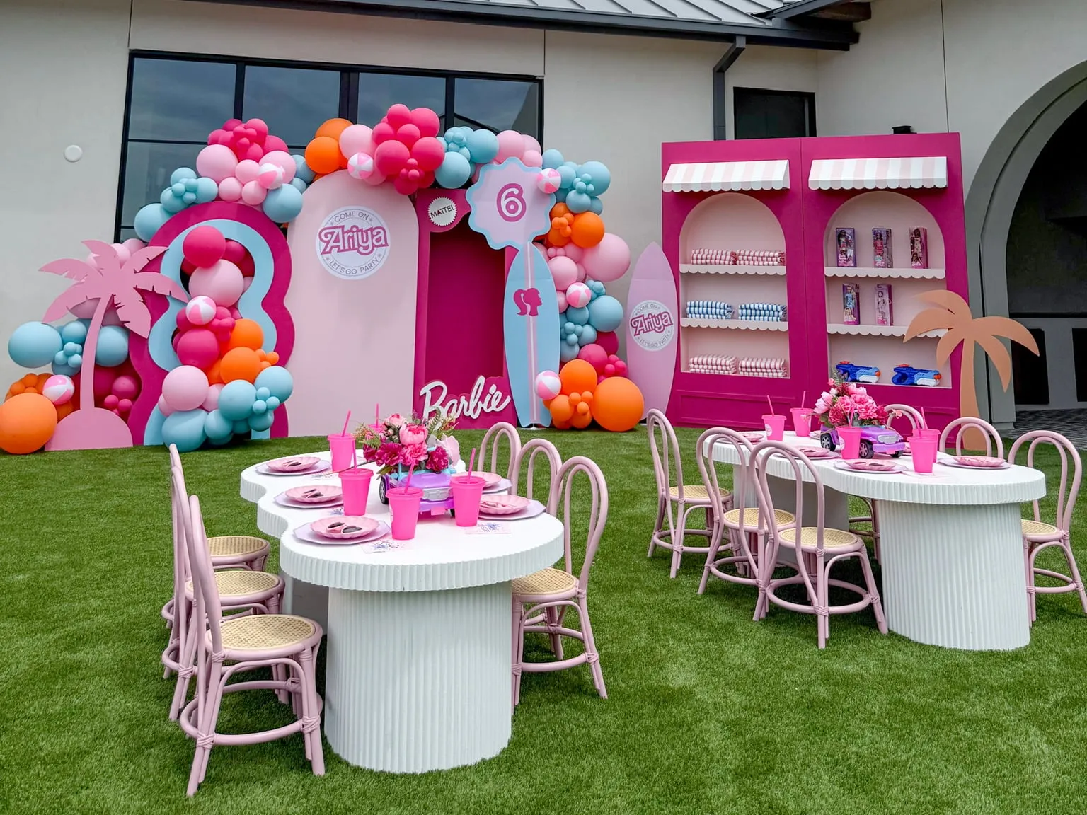 Barbie themed sixth birthday with pink and blue arch backdrops, a surfboard, a pink storefront shelf display, balloon garland and styled kids tables on the lawn
