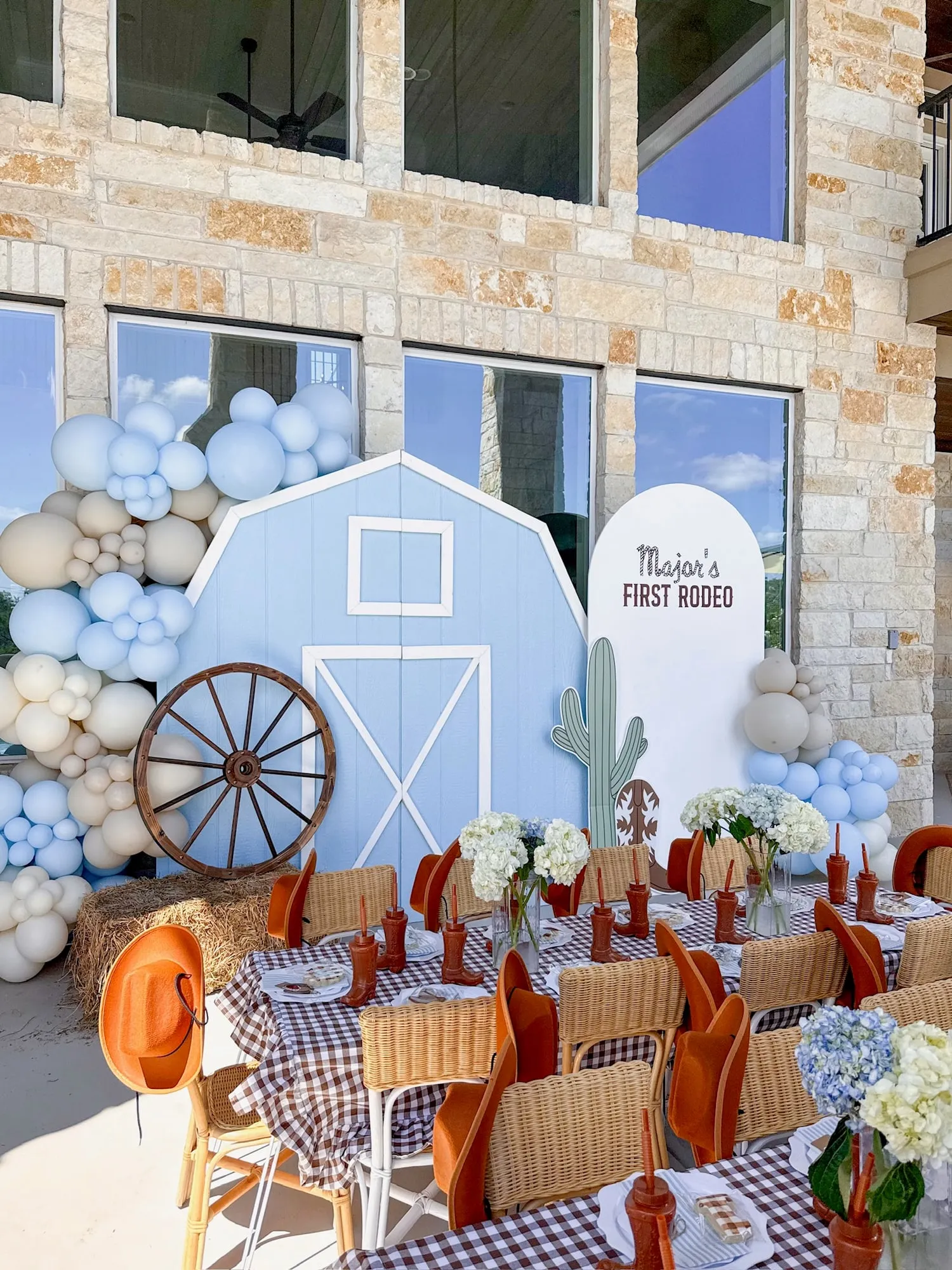 First rodeo birthday with a blue barn backdrop, wagon wheel, blue and sand balloon garland and gingham kids tables with cowboy hats