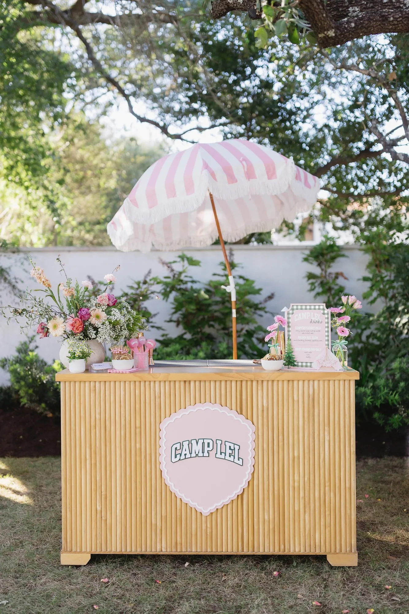 Fluted wood bar with a pink striped fringe umbrella, custom camp sign, drink menu and florals