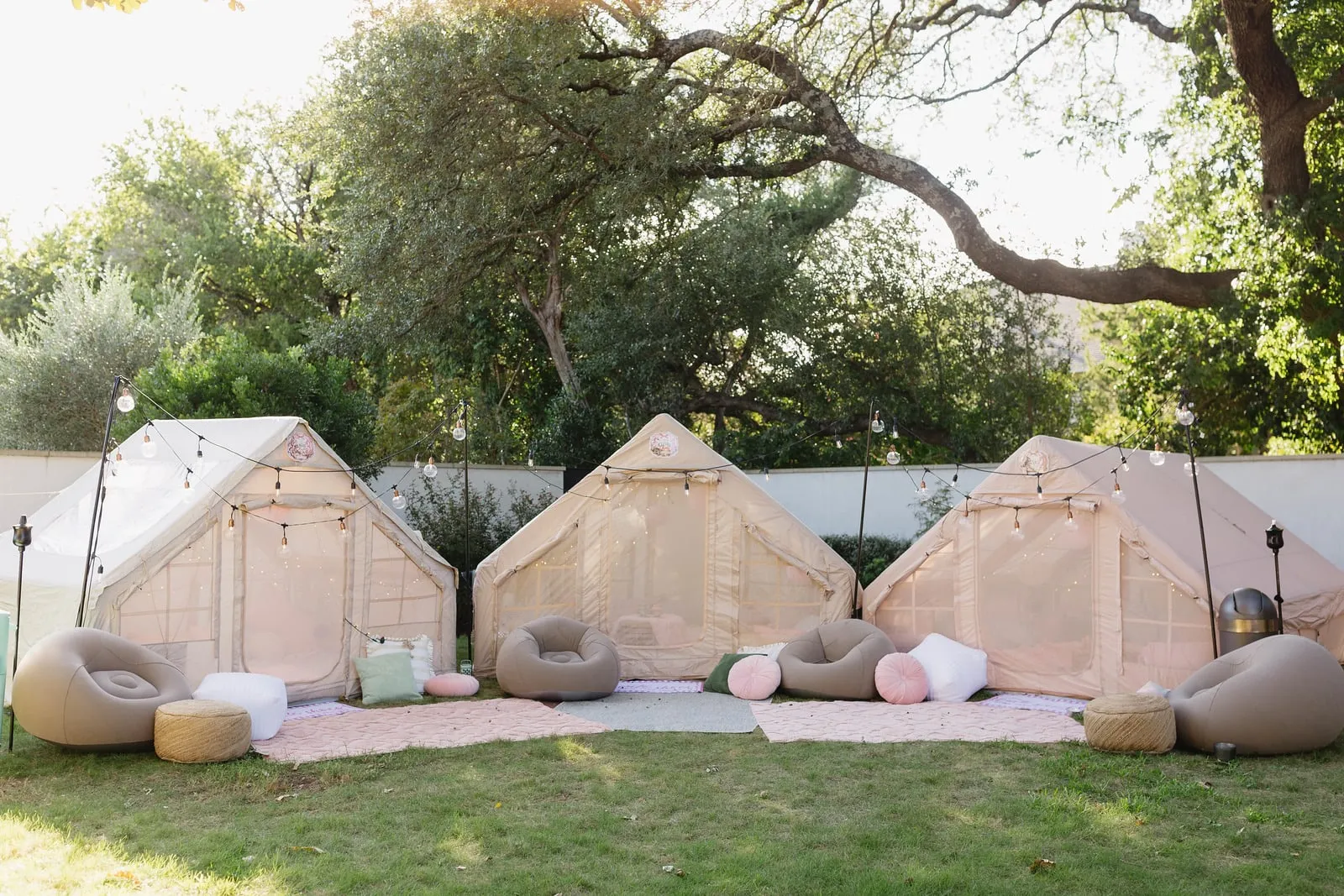 Three glamping tents with string lights, poufs and pink rugs set up for a backyard sleepover