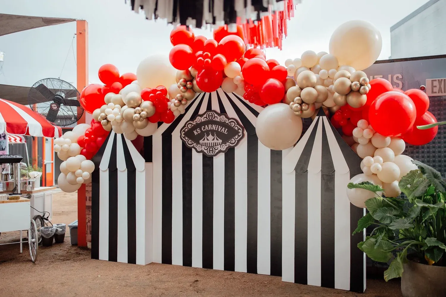 Black and white striped carnival tent backdrop with red, cream and gold balloon garland for Kendra Scott