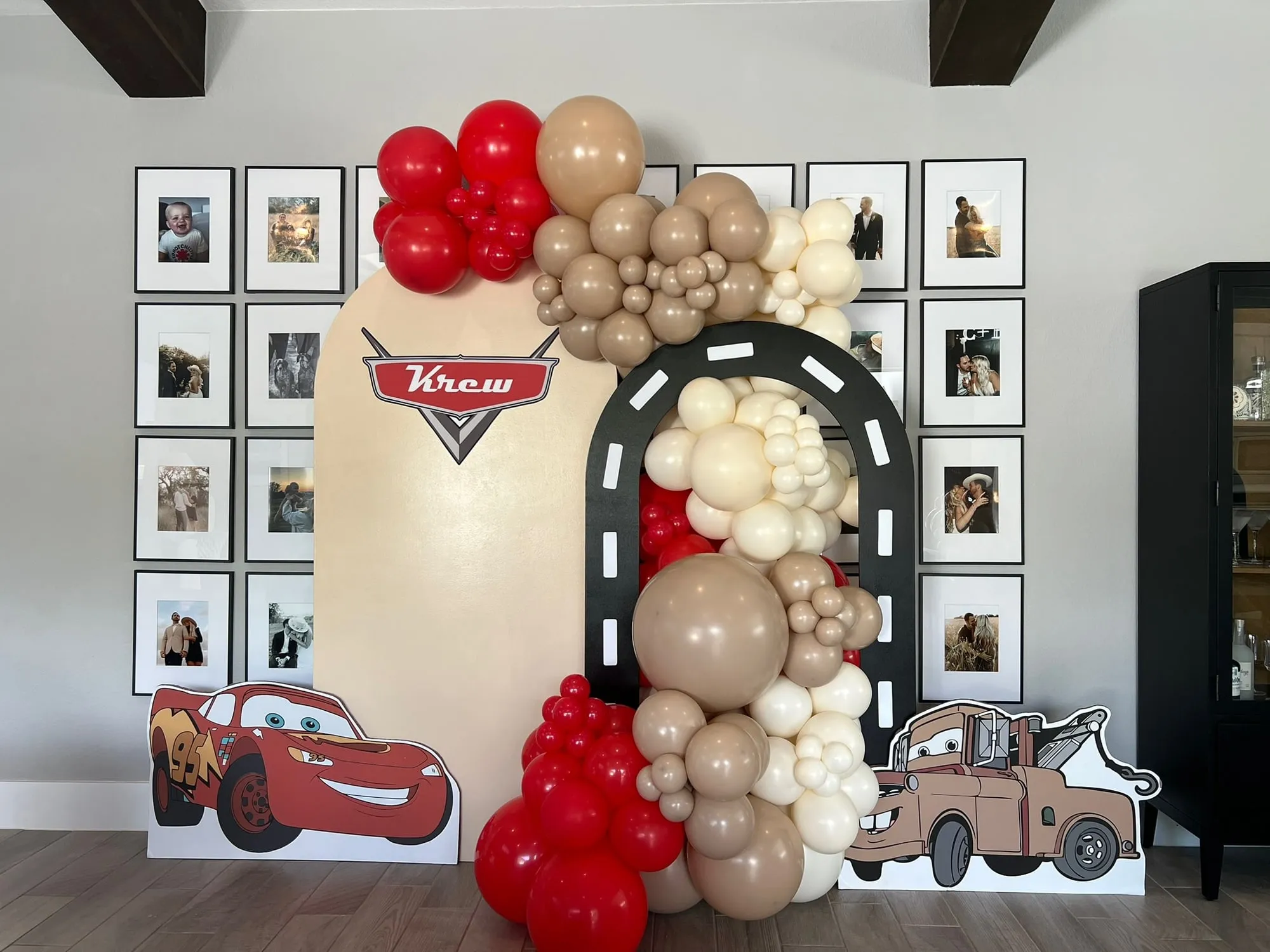 Cars themed backdrop with a racetrack arch, Lightning McQueen and Mater cutouts and a red, tan and cream balloon garland