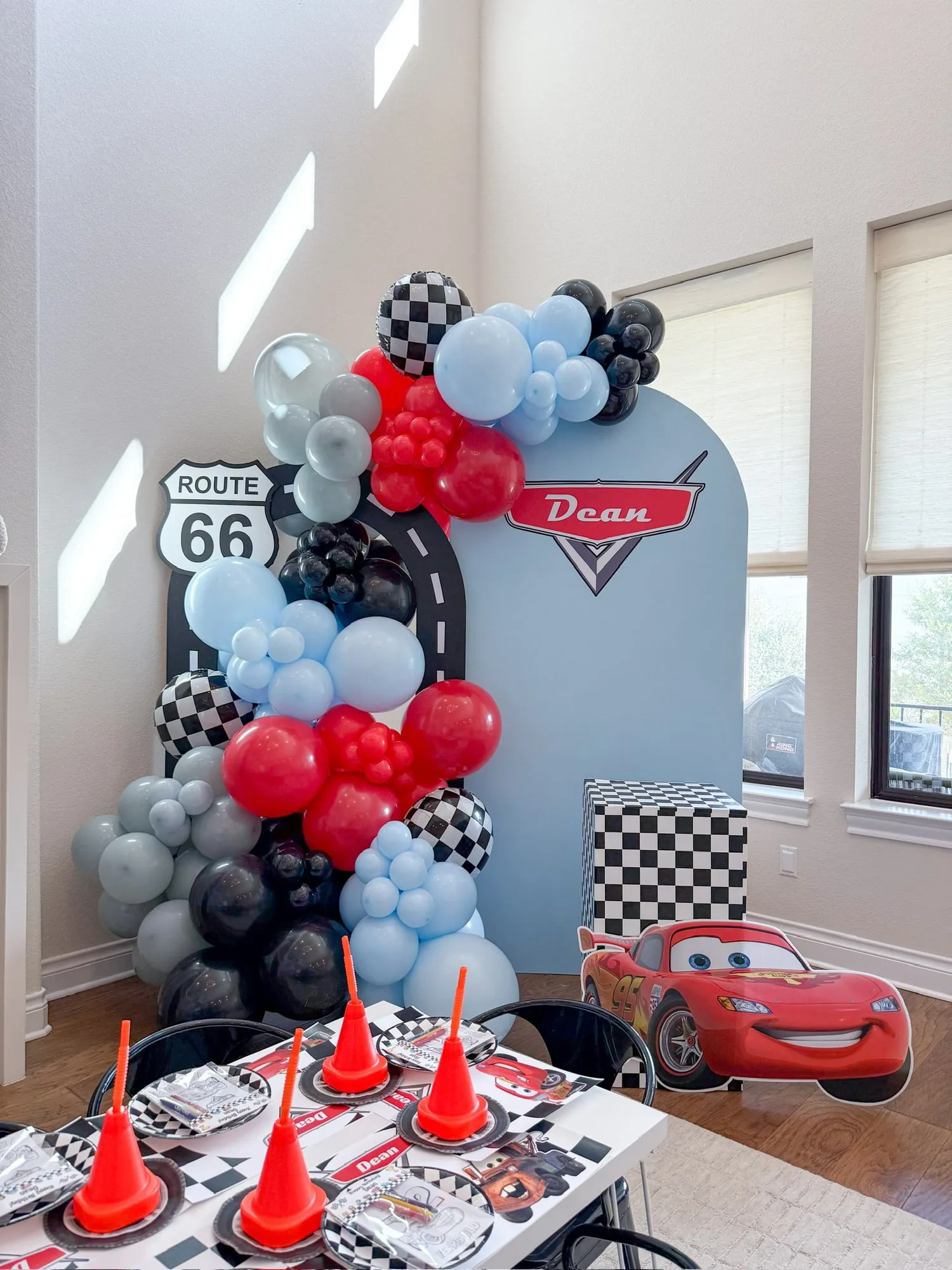 Cars themed birthday with a blue arch backdrop, Route 66 sign, checkered pedestal and red, blue and black balloon garland