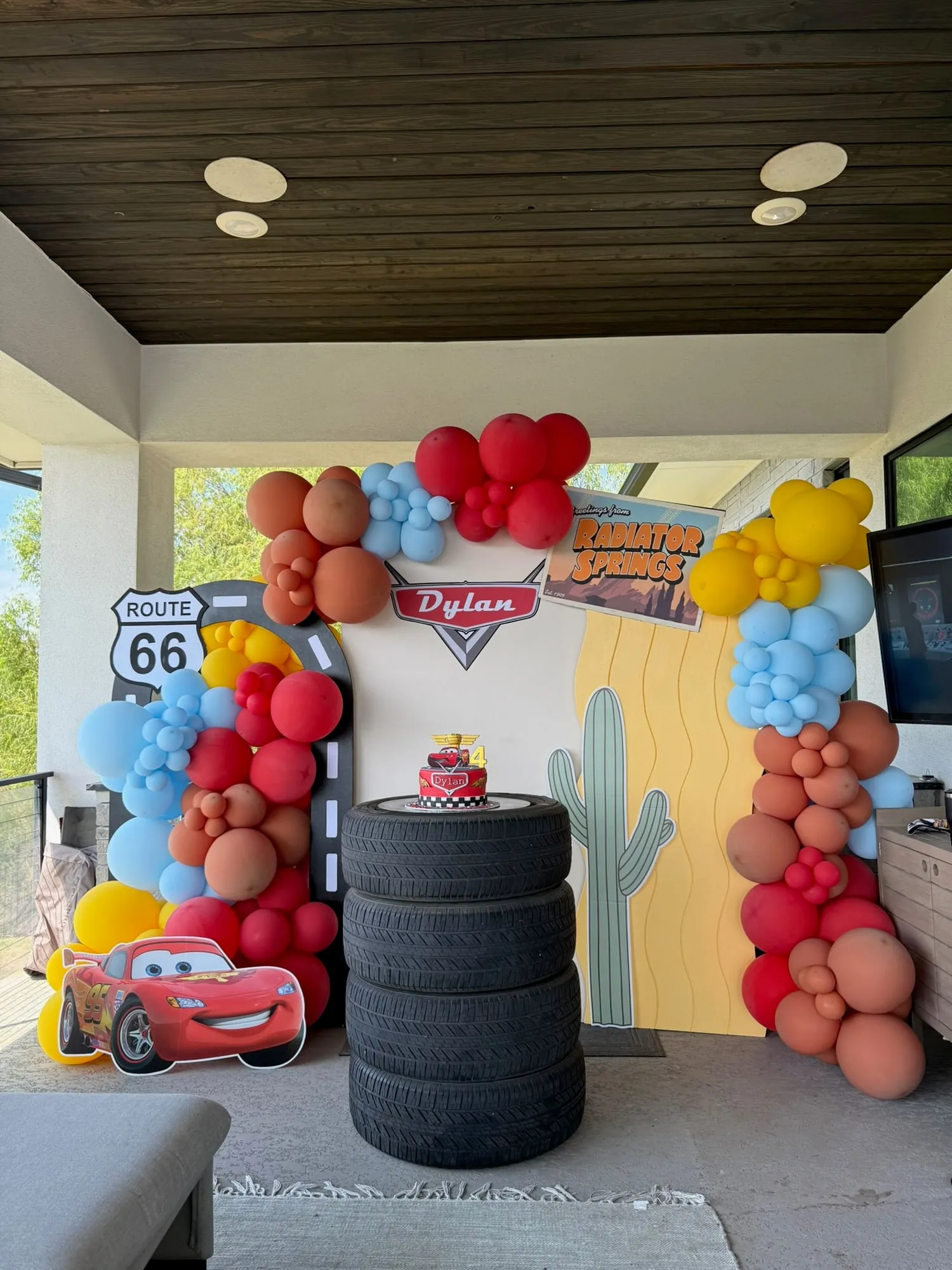 Cars themed fourth birthday with a Route 66 road arch, Radiator Springs backdrop, tire cake stand and red, rust and blue balloon garland