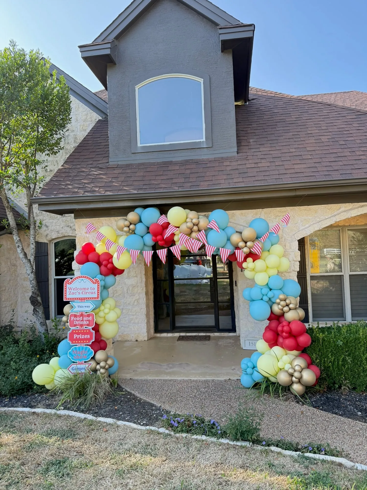 Circus themed balloon arch in red, aqua, yellow and gold with striped pennants and a welcome sign framing a front door