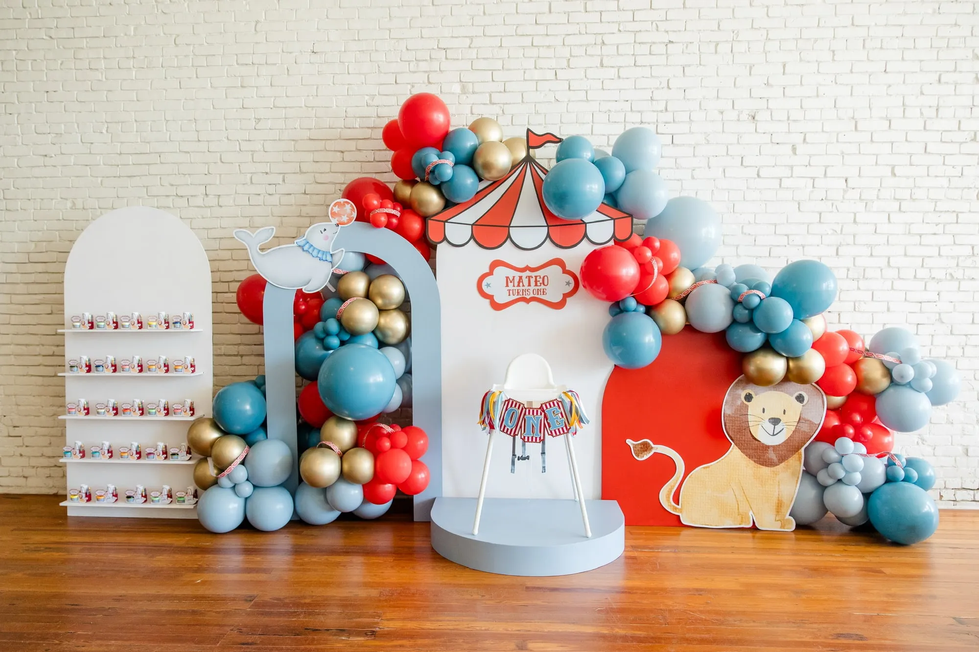 Circus themed first birthday with a big top panel, lion and seal cutouts, a favor shelf and red, blue and gold balloon garland