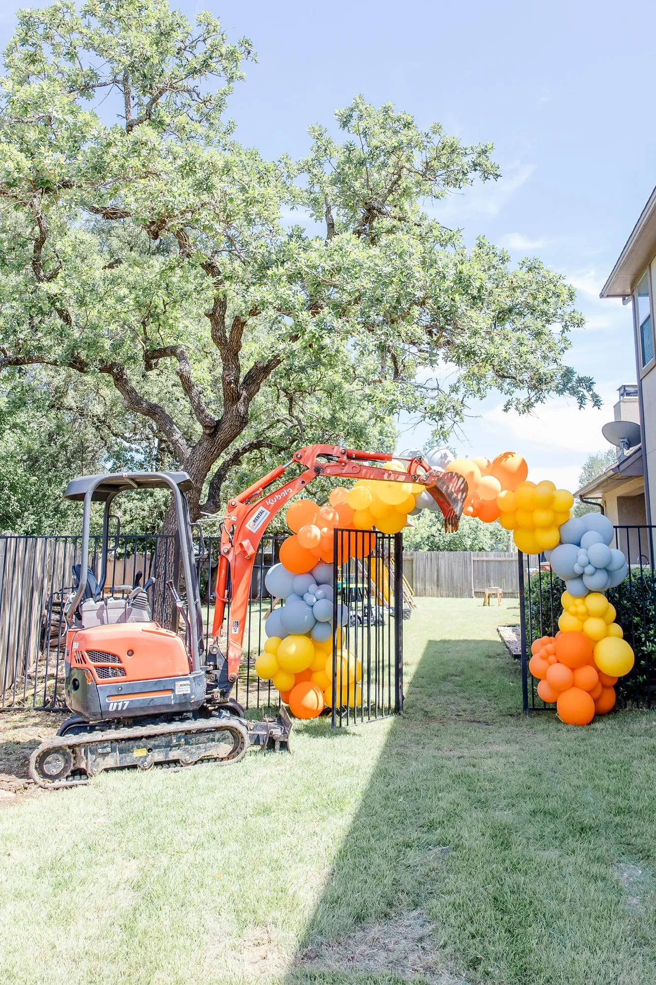 Construction themed backyard balloon arch in orange, yellow and slate held up by a real mini excavator