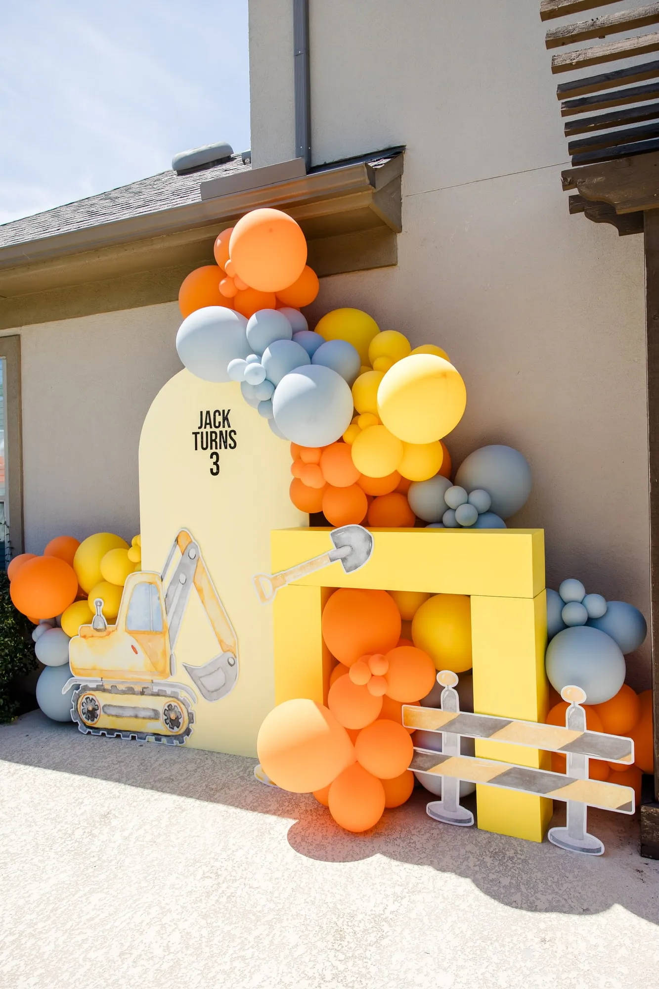 Construction themed third birthday with a yellow arch backdrop, excavator cutout, road barrier and orange, yellow and slate balloon garland