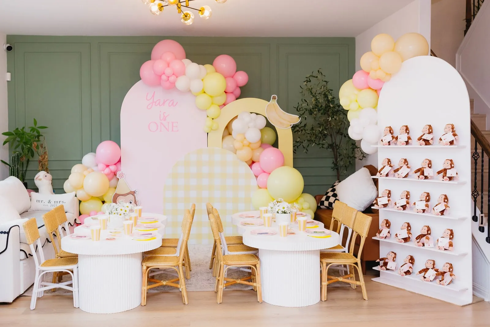 Curious George first birthday with pink and yellow arch backdrops, gingham panel, pastel balloon garlands and styled kids tables