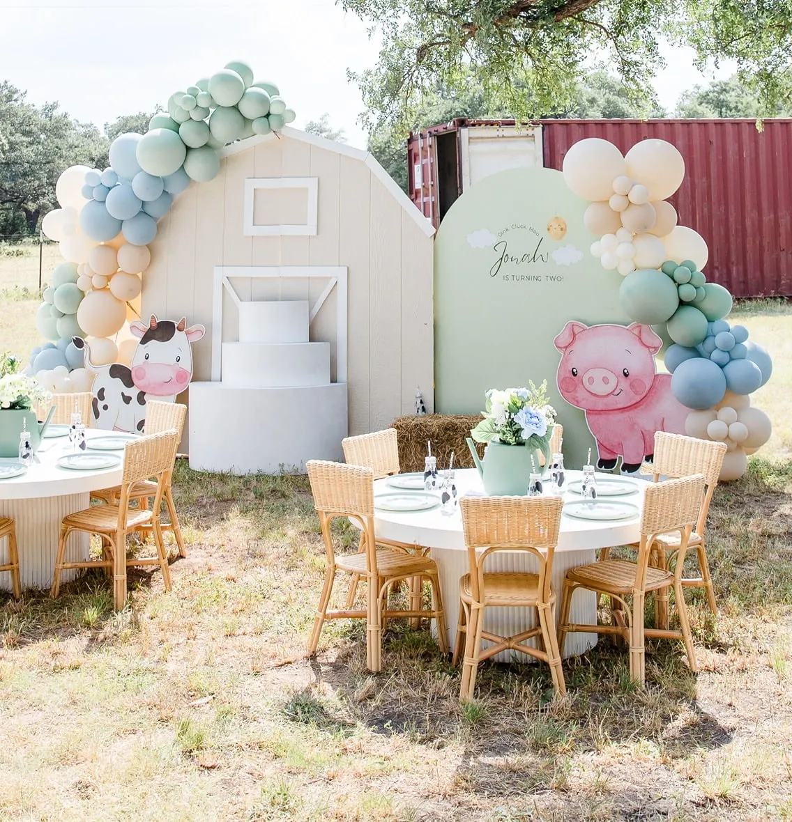 Farm themed second birthday with a cream barn backdrop, sage, blue and cream balloon garland, pig and cow cutouts and rattan kids tables outdoors
