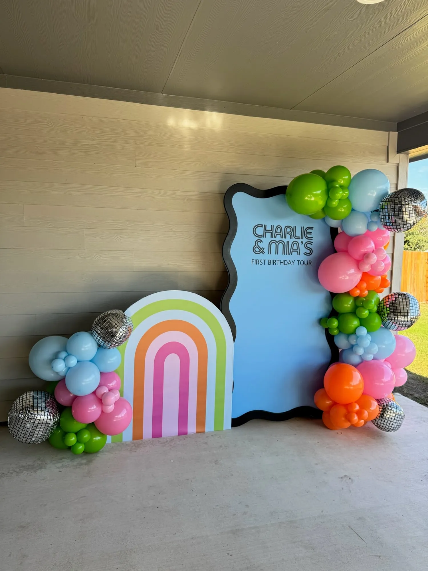 First Birthday Tour twin party backdrop with a rainbow arch, blue wavy panel and bright balloon garland with disco ball balloons