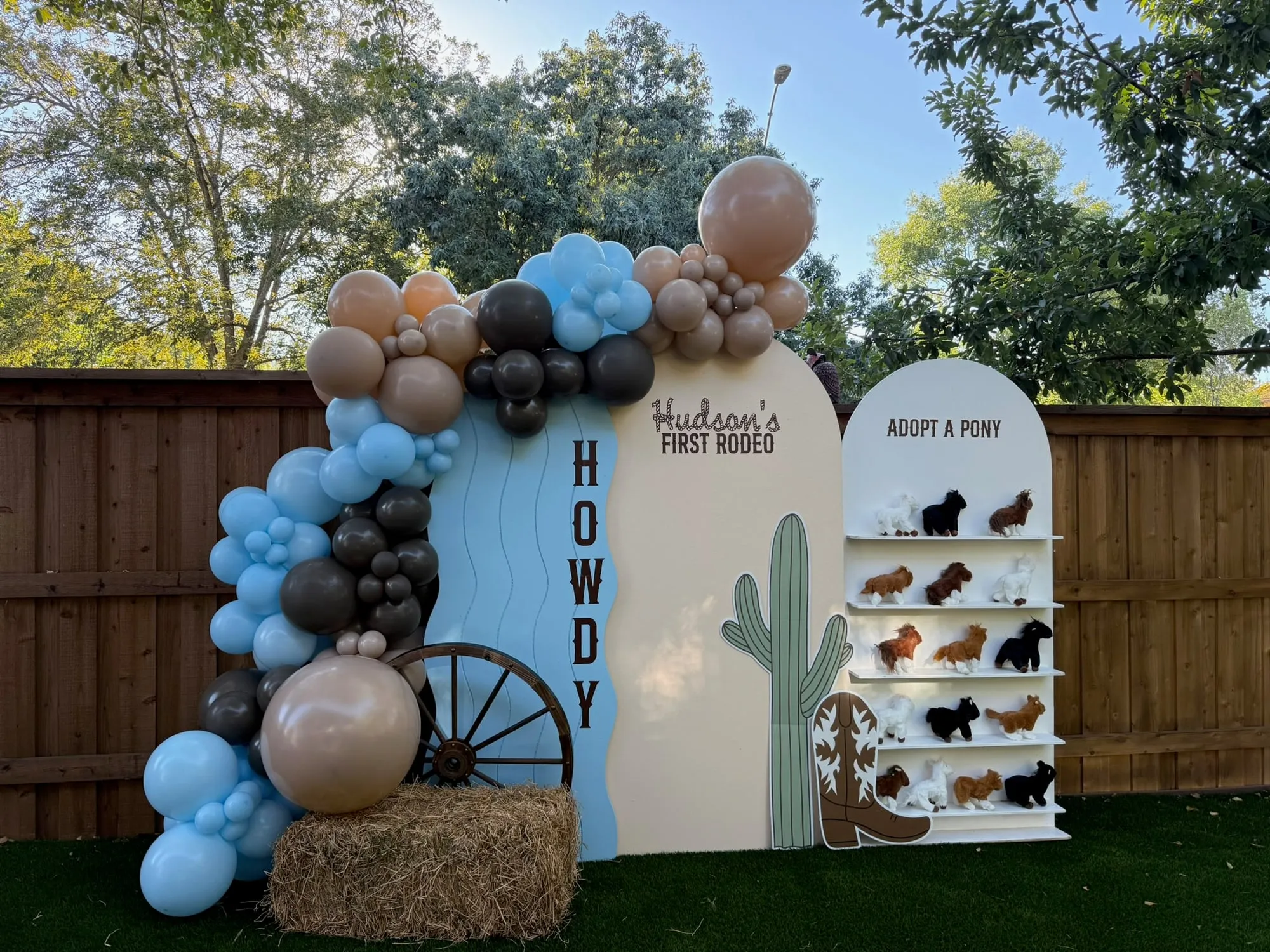 First rodeo birthday with a blue Howdy panel, cactus and boot cutouts, wagon wheel, adopt a pony shelf and blue and tan balloon garland