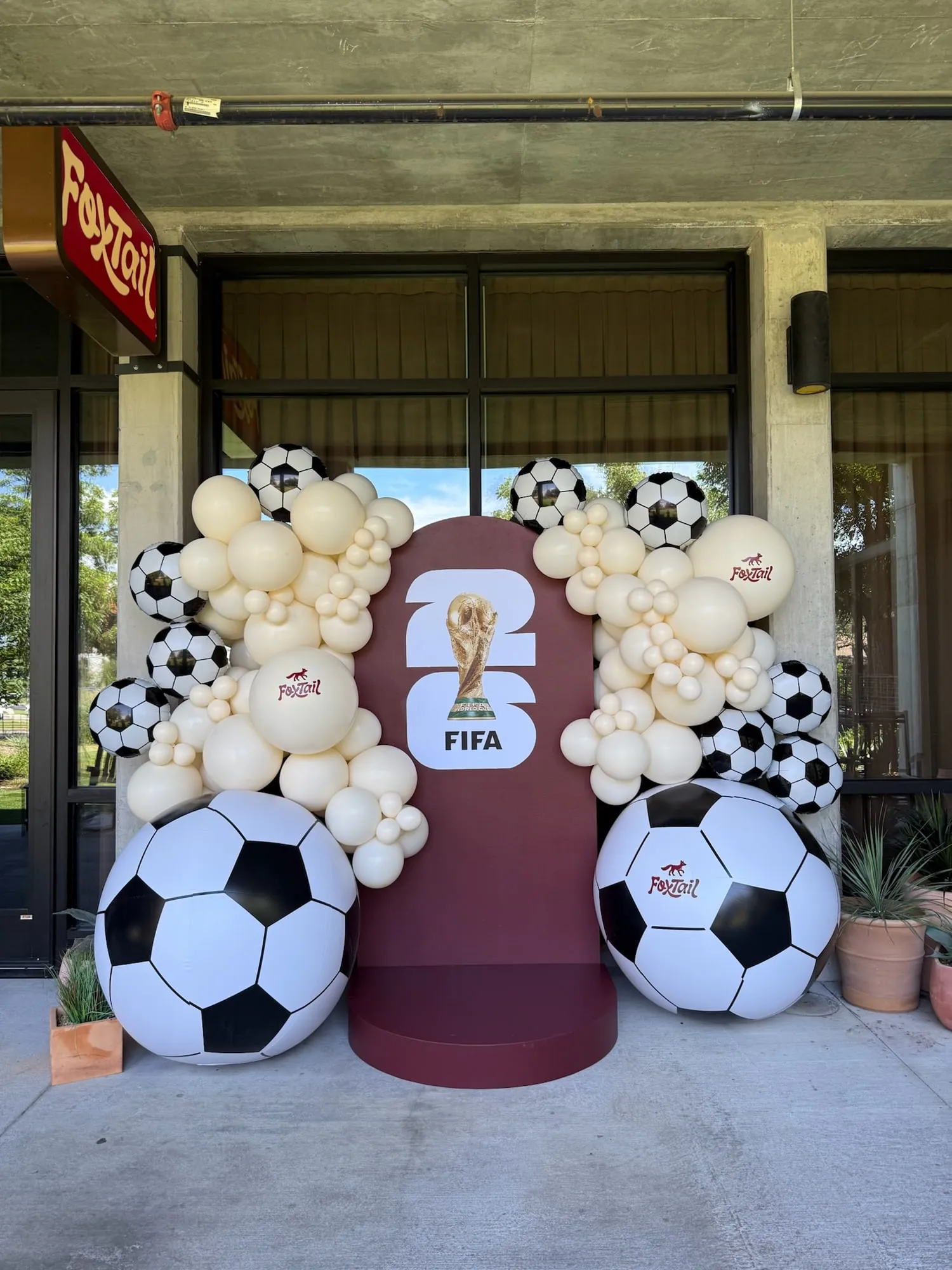 Soccer watch party backdrop at Foxtail with a burgundy arch, cream balloon garland, branded balloons and giant soccer balls