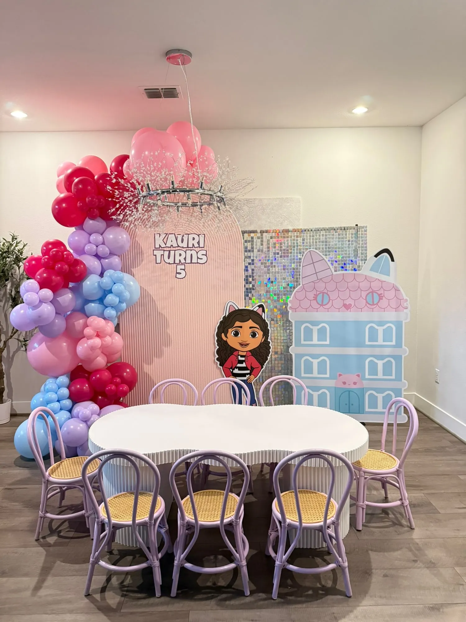 Gabby’s Dollhouse themed fifth birthday with a pink fluted arch, dollhouse cutout, shimmer wall and a cloud kids table with lilac chairs