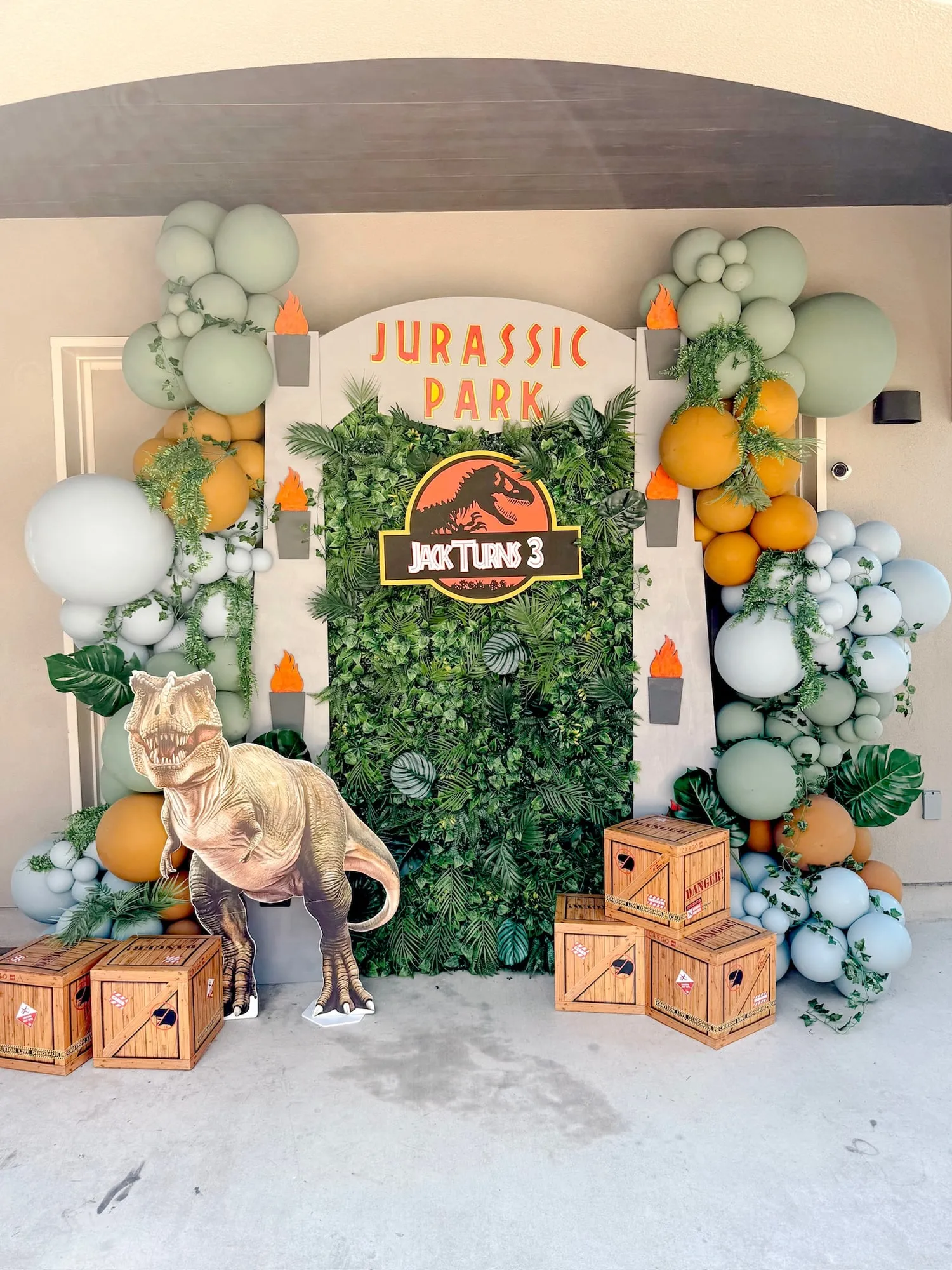Jurassic Park themed third birthday with a gate backdrop, greenery wall, sage, mustard and blue balloon garland and a T. rex cutout