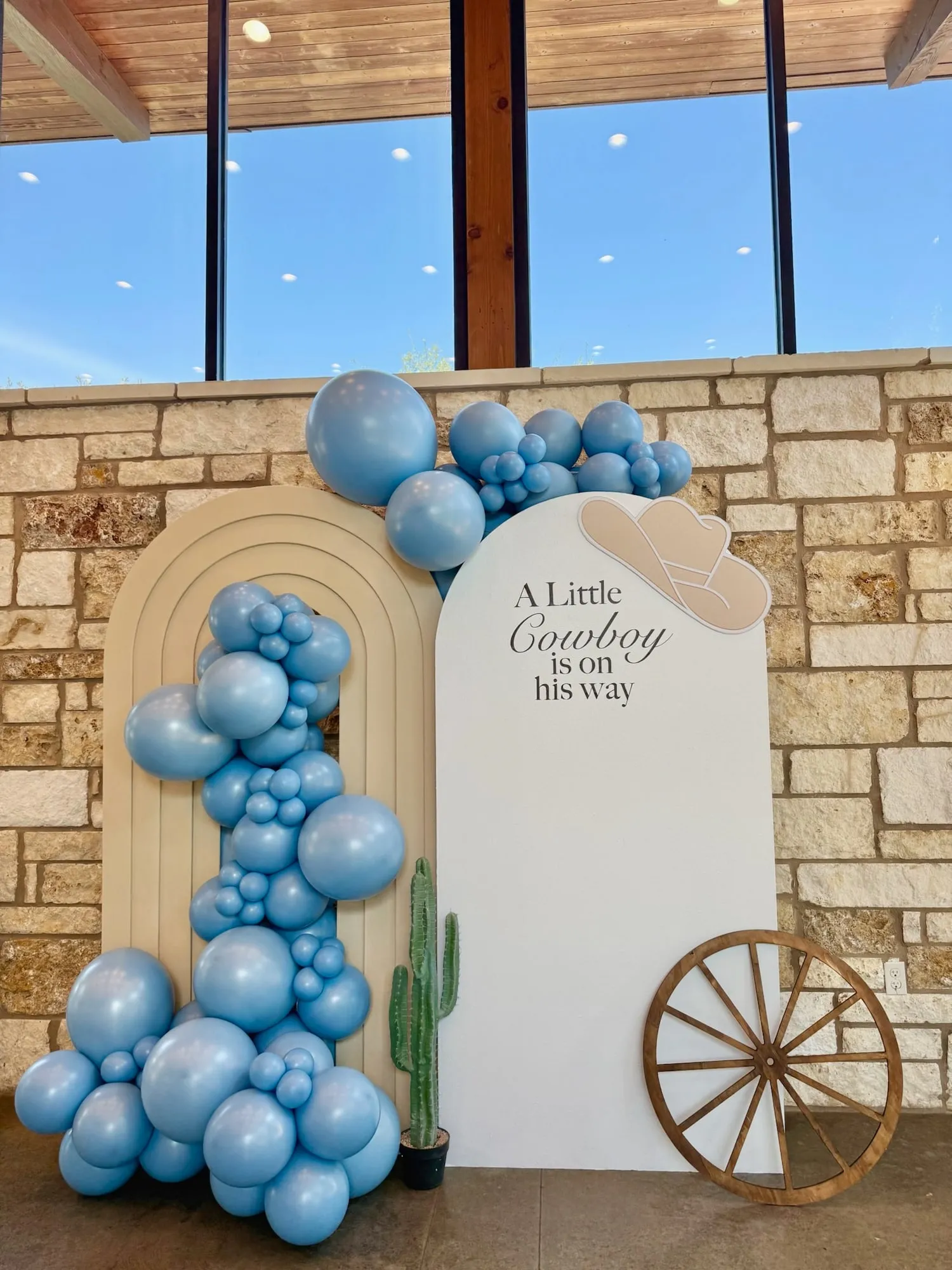 Little cowboy baby shower with a tan rainbow arch, white arch with cowboy hat, dusty blue balloon garland, cactus and wagon wheel