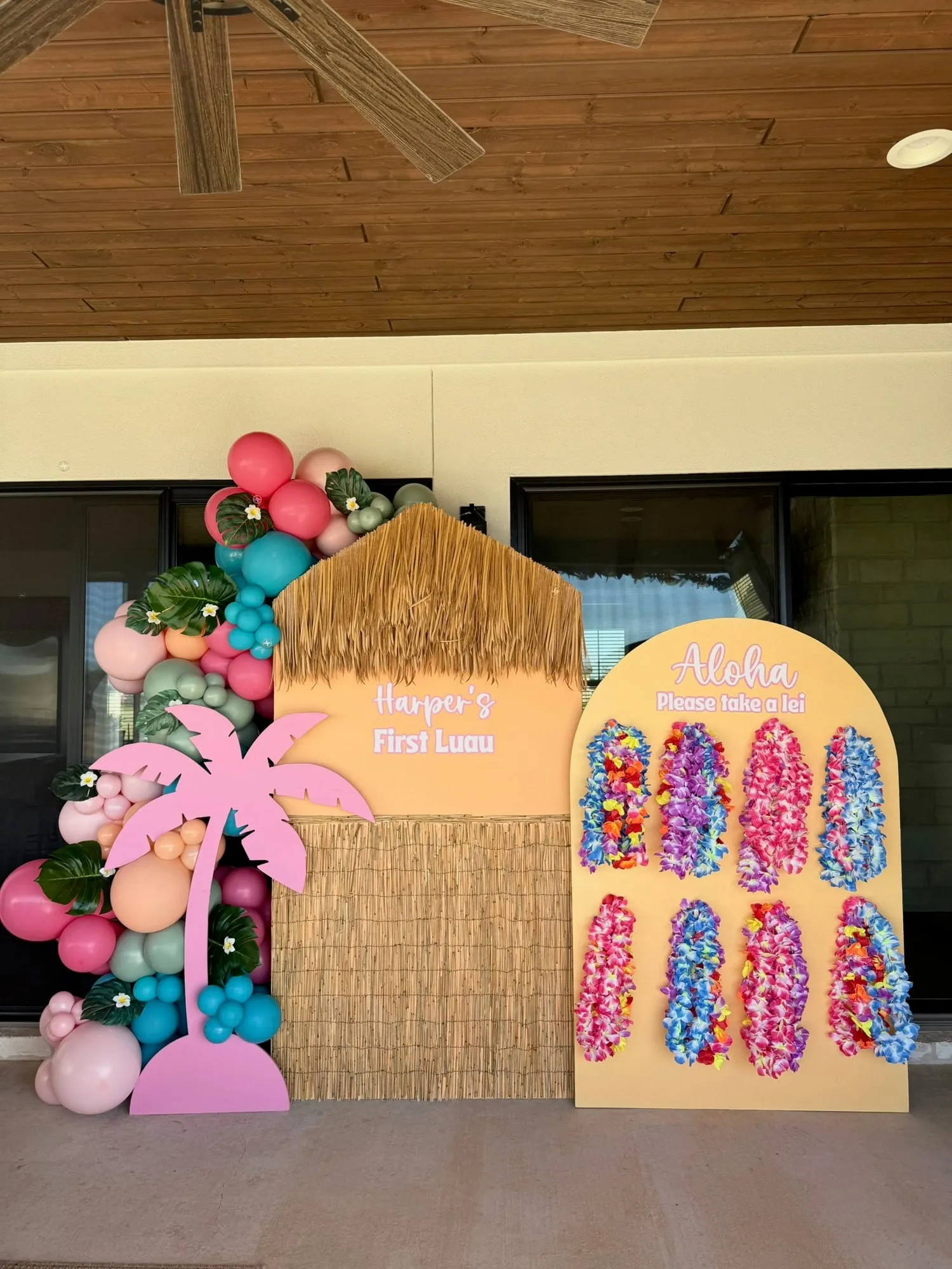 Luau first birthday with a thatched tiki hut backdrop, pink palm tree, lei wall and tropical balloon garland