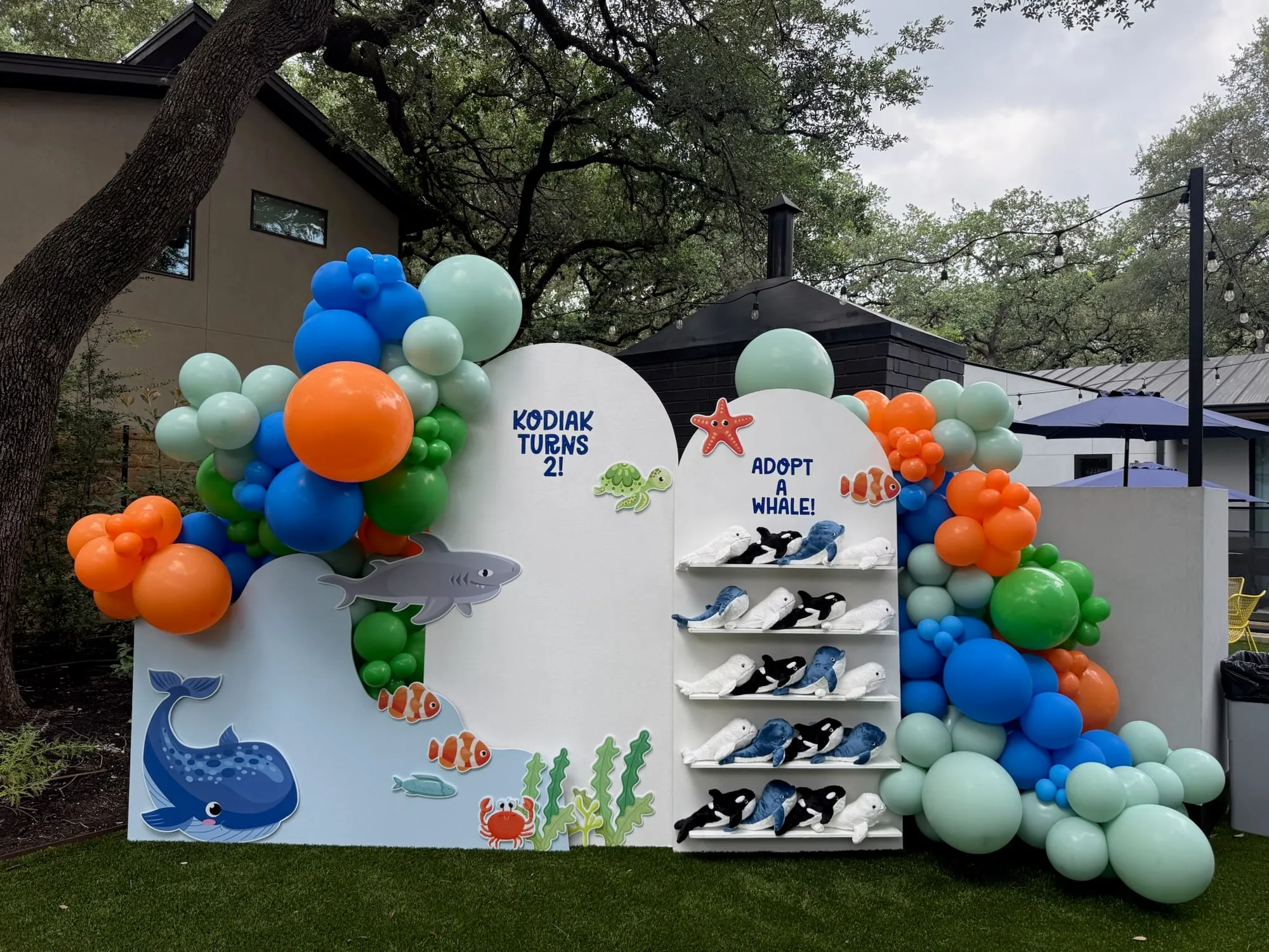 Ocean themed second birthday with a wave backdrop, sea creature cutouts, an adopt a whale plush shelf and blue, orange and green balloons