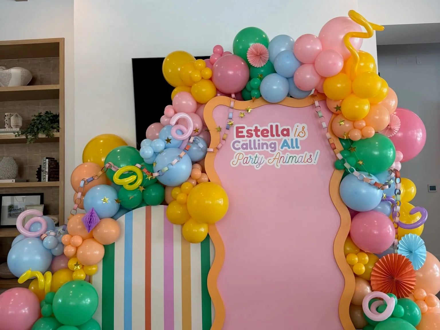 Party animals birthday backdrop with wavy pink and orange panel, rainbow striped arch and colorful balloon garland