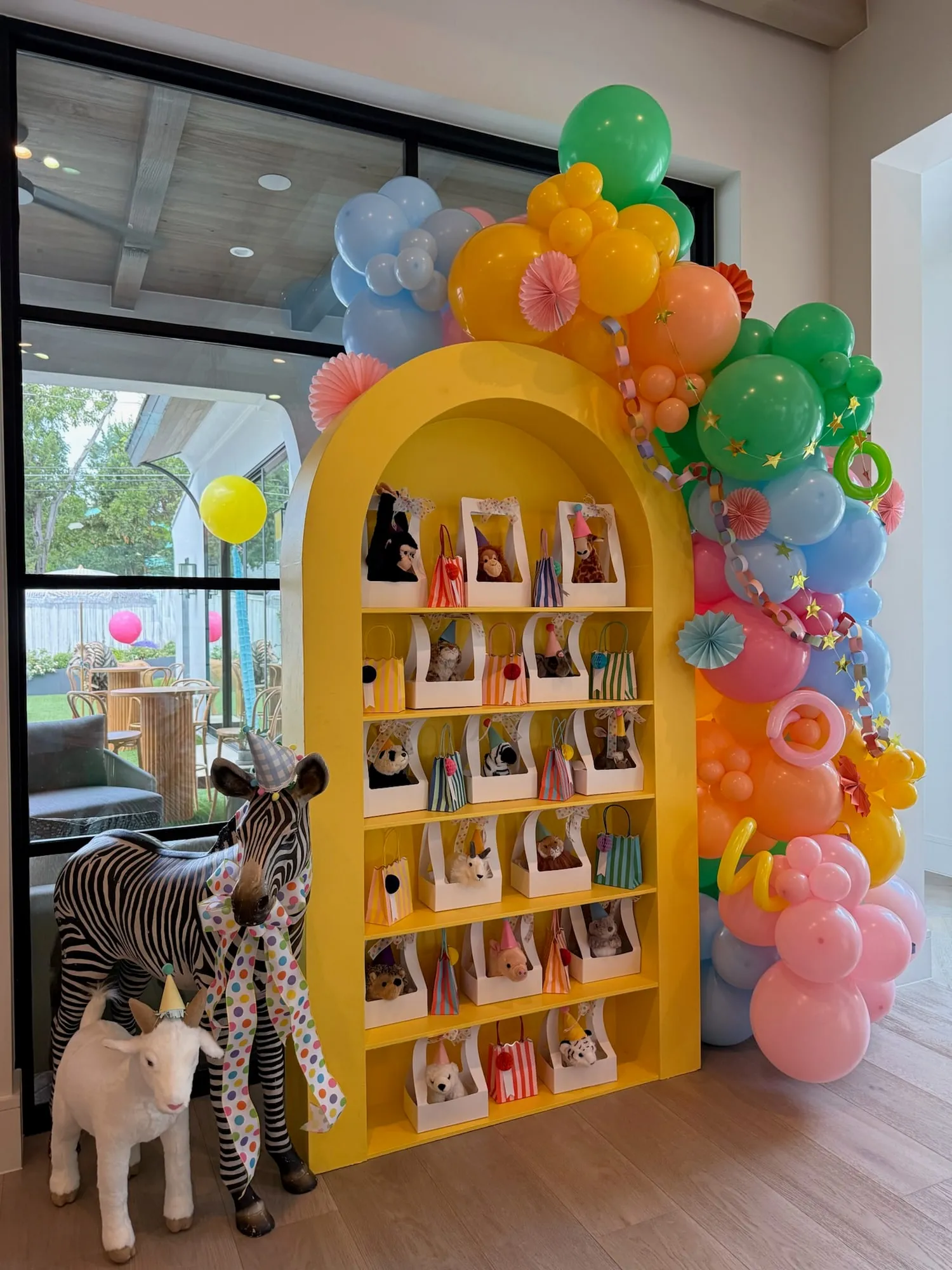 Yellow arch favor shelf with plush animals in party hats and a rainbow balloon garland with paper chains and fans