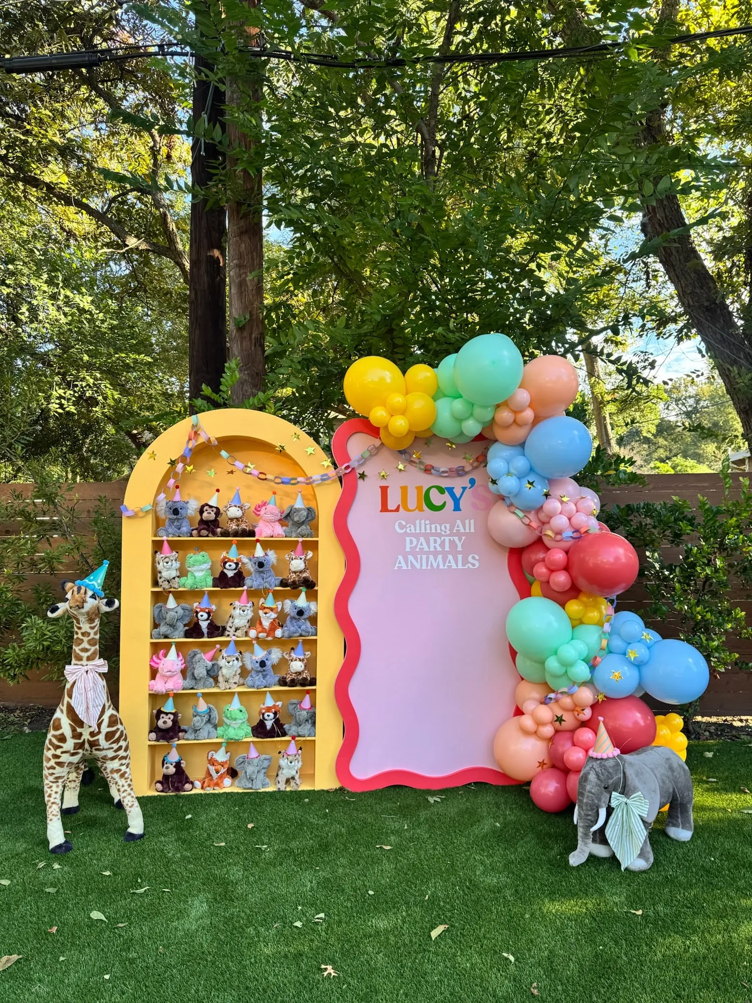 Calling All Party Animals first birthday with a yellow plush animal shelf, red wavy panel and rainbow balloon garland