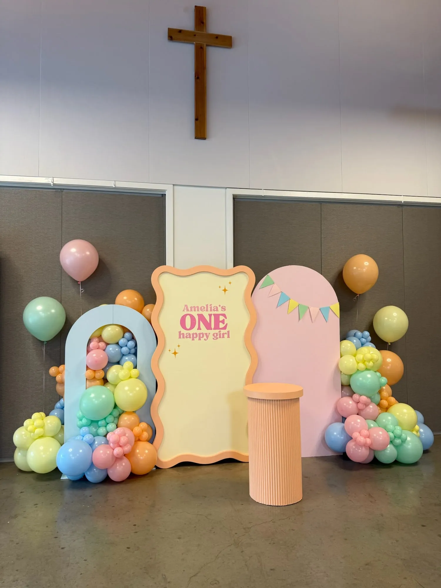 Pastel first birthday in a church hall with blue, cream wavy and pink arch backdrops, a fluted pedestal and rainbow balloon clusters