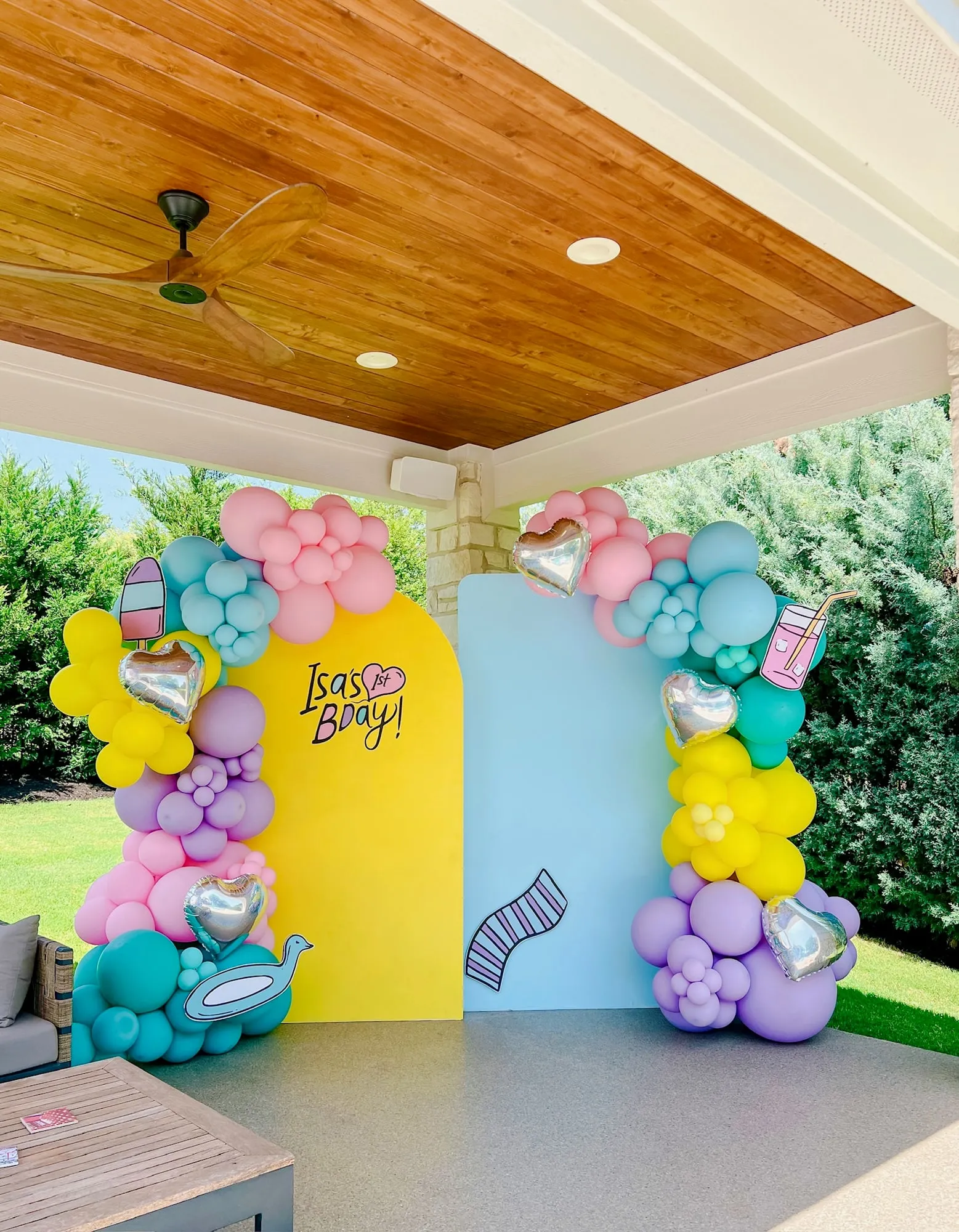 Pastel summer first birthday with yellow and blue backdrops, rainbow balloon garland and silver heart balloons on a covered patio