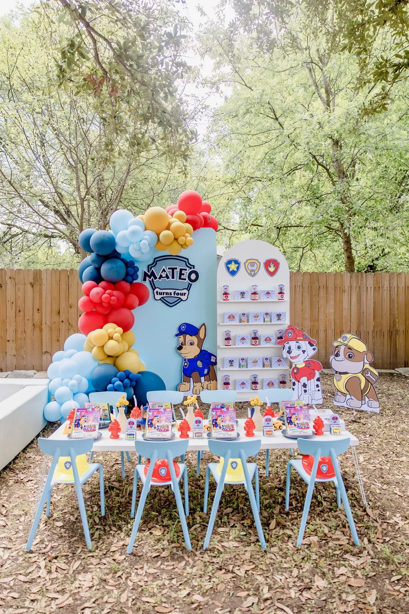 Paw Patrol themed fourth birthday with blue backdrop, red, yellow and blue balloon garland, favor wall and styled kids table in a backyard