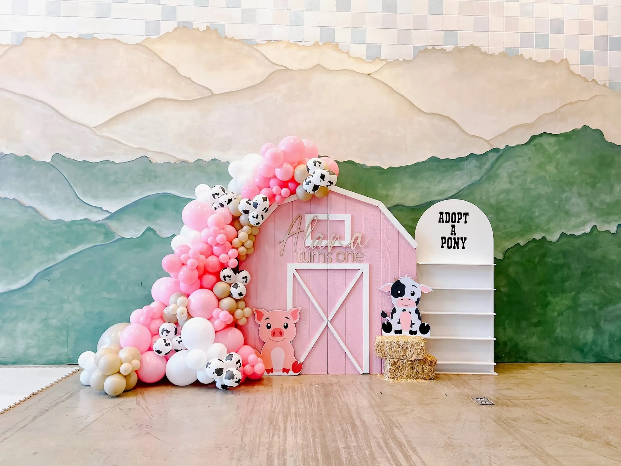 Pink barn farm themed first birthday backdrop with pink, white and cow print balloon garland, pig and cow cutouts and a favor shelf