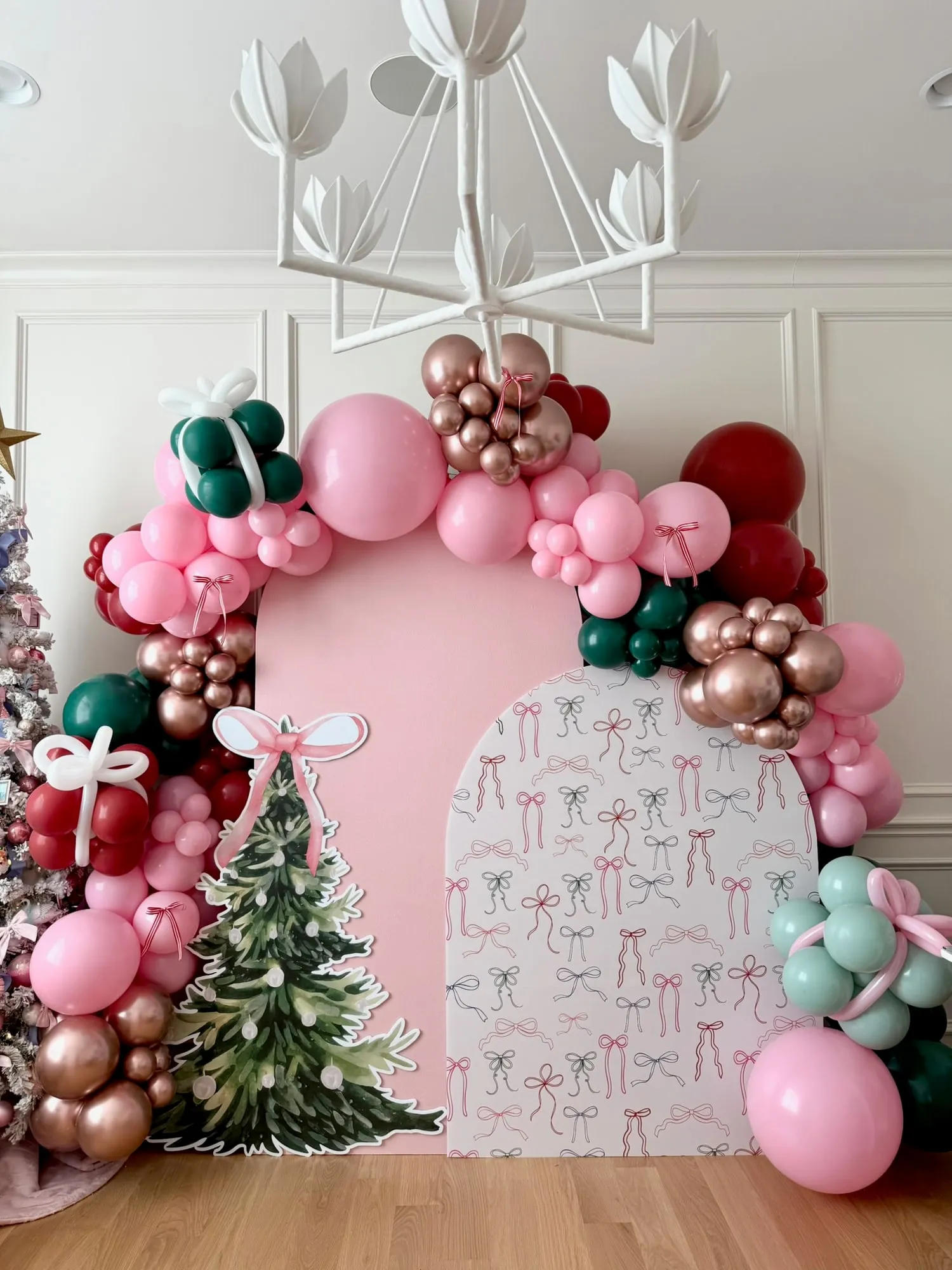 Pink Christmas backdrop with a bow print arch, watercolor tree cutout and pink, rose gold, burgundy and green balloon garland with present balloons