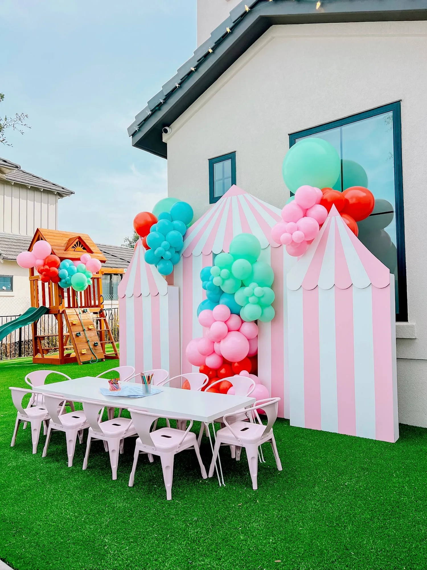 Pink and white striped circus tent backdrops with pink, teal and red balloon clusters and a kids table on the lawn