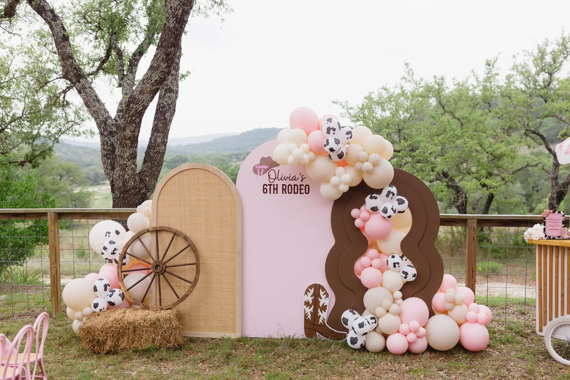 Pink cowgirl sixth birthday with rattan and pink arches, a wavy brown panel, wagon wheel and cow print balloon garland overlooking the Hill Country