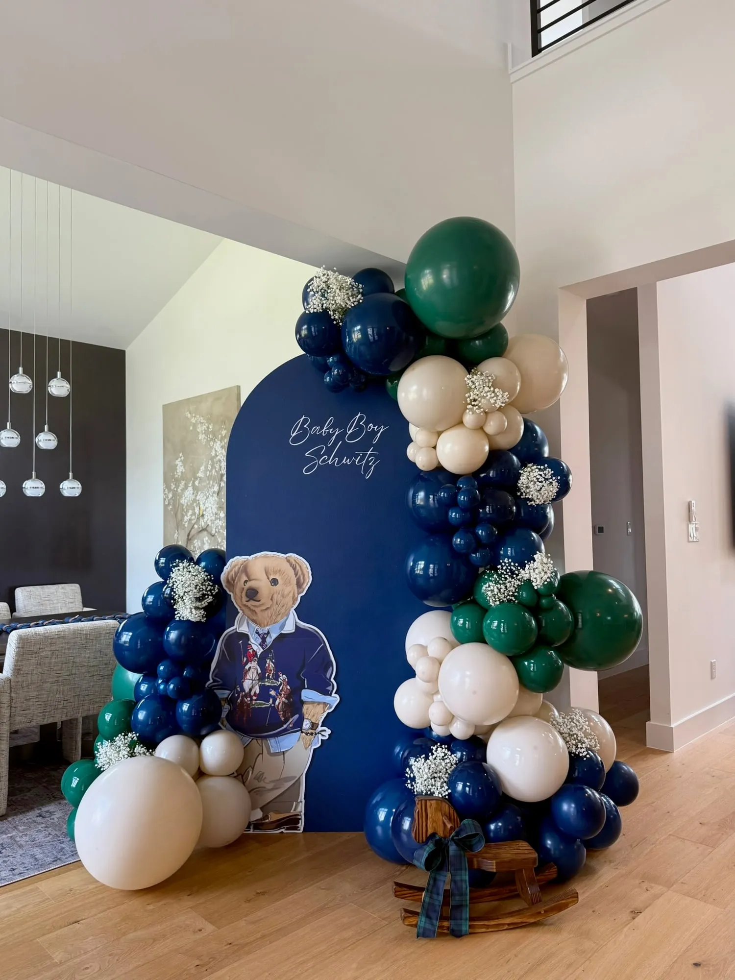 Navy arch baby shower backdrop with a preppy bear cutout, navy, green and cream balloon garland, baby’s breath and a rocking horse