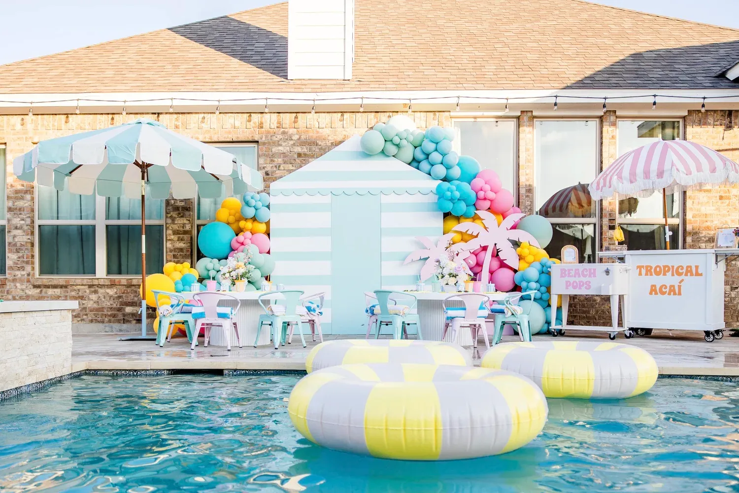 Beach themed pool party with pastel balloon garland, striped cabana backdrop, kids tables and snack carts