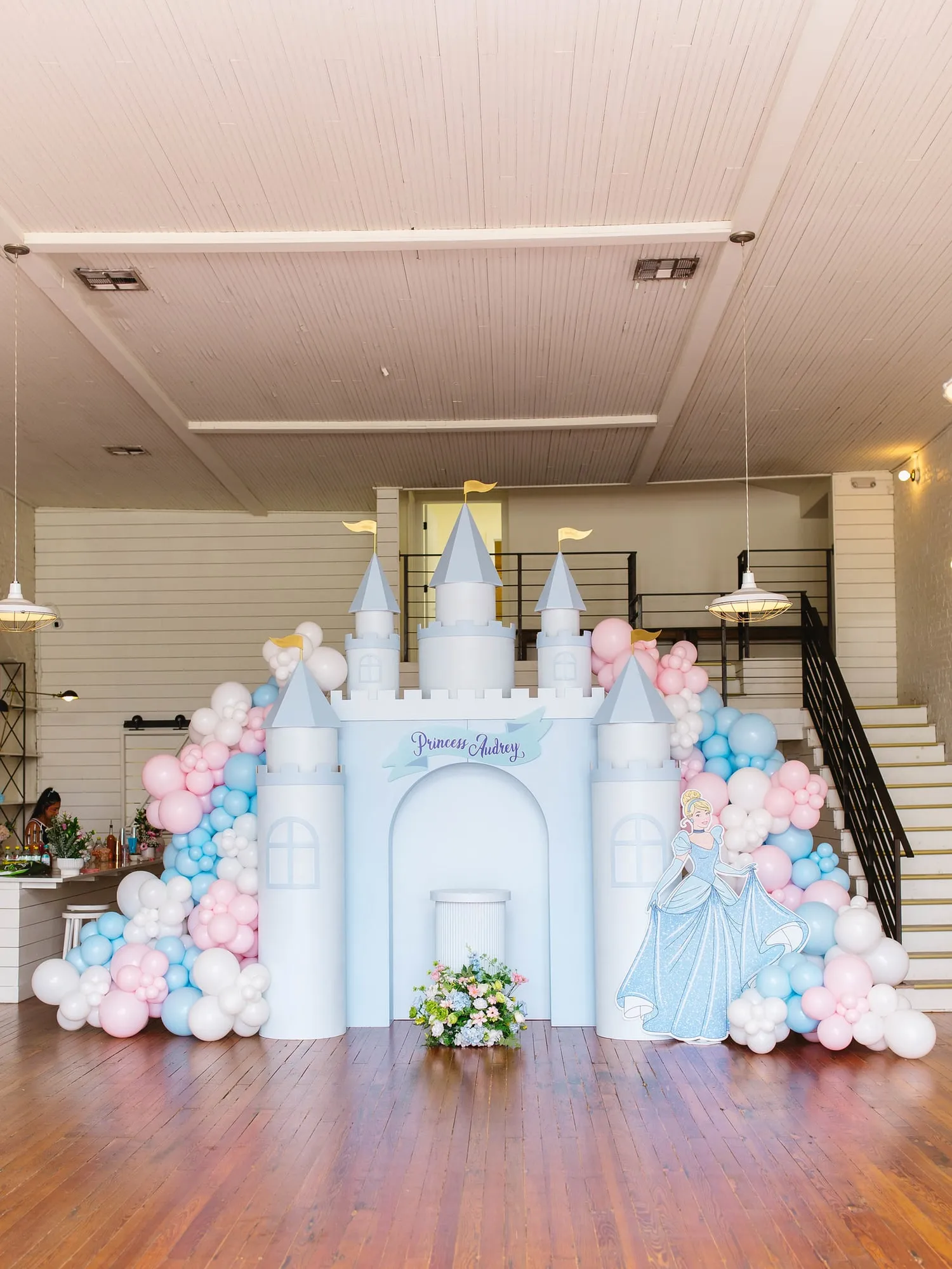 Princess castle backdrop with turrets and gold flags, pink, blue and white balloon garland and a floral arrangement in an event hall
