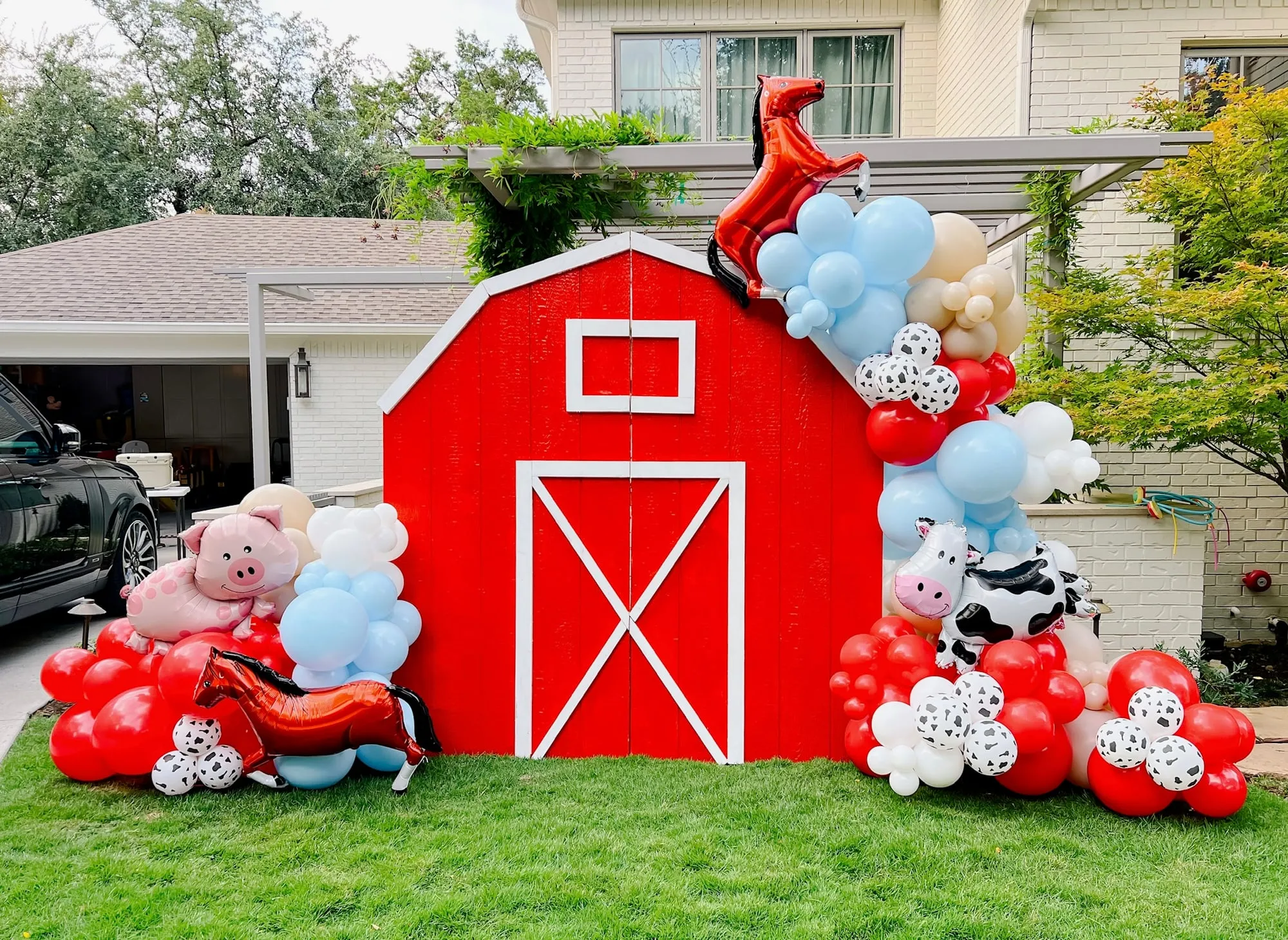 Red barn backdrop with red, blue and cow print balloon garland and horse, pig and cow foil balloons on a front lawn
