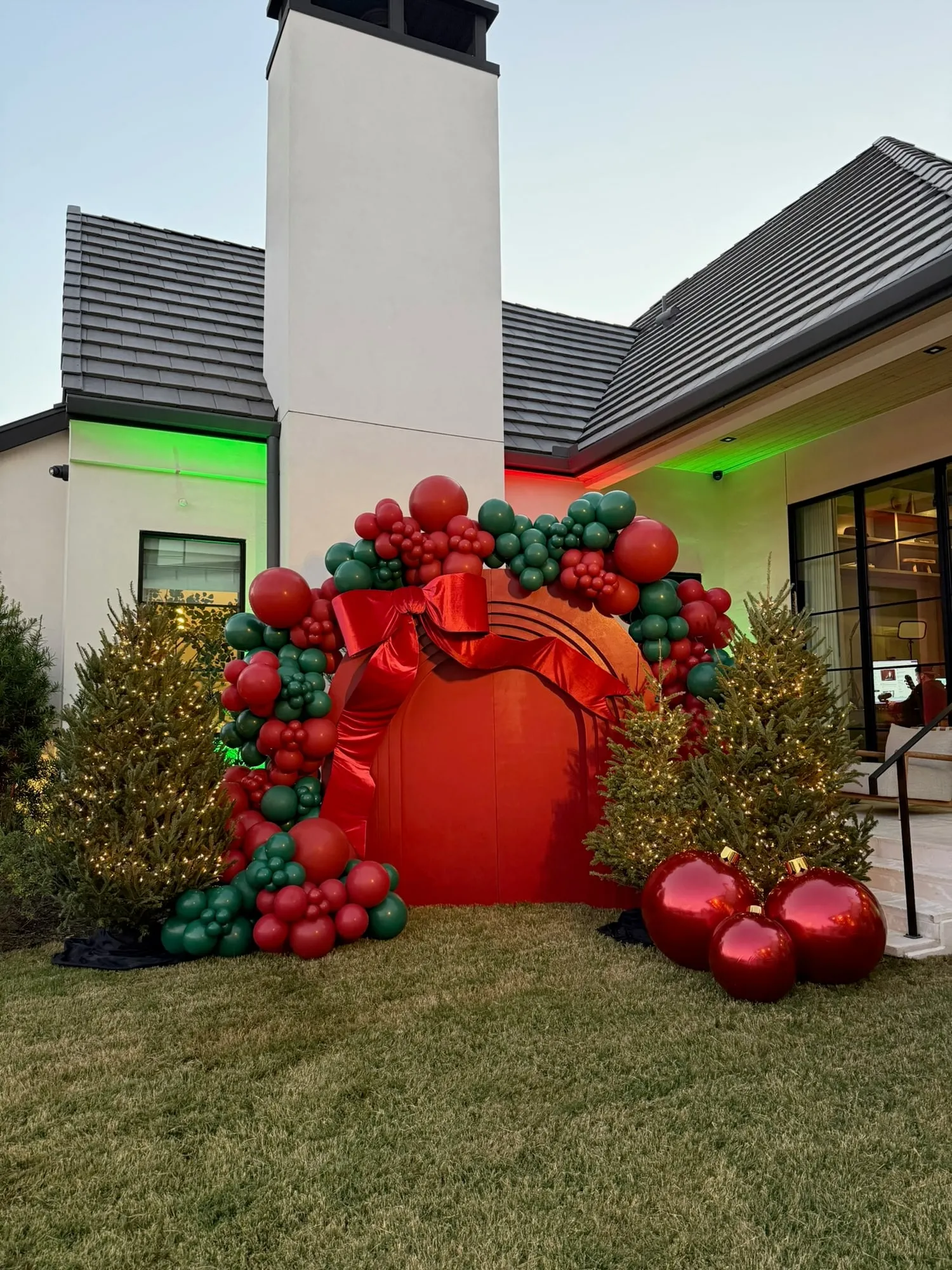 Red Christmas arch backdrop with an oversized velvet bow, red and green balloon garland, lit trees and giant ornaments