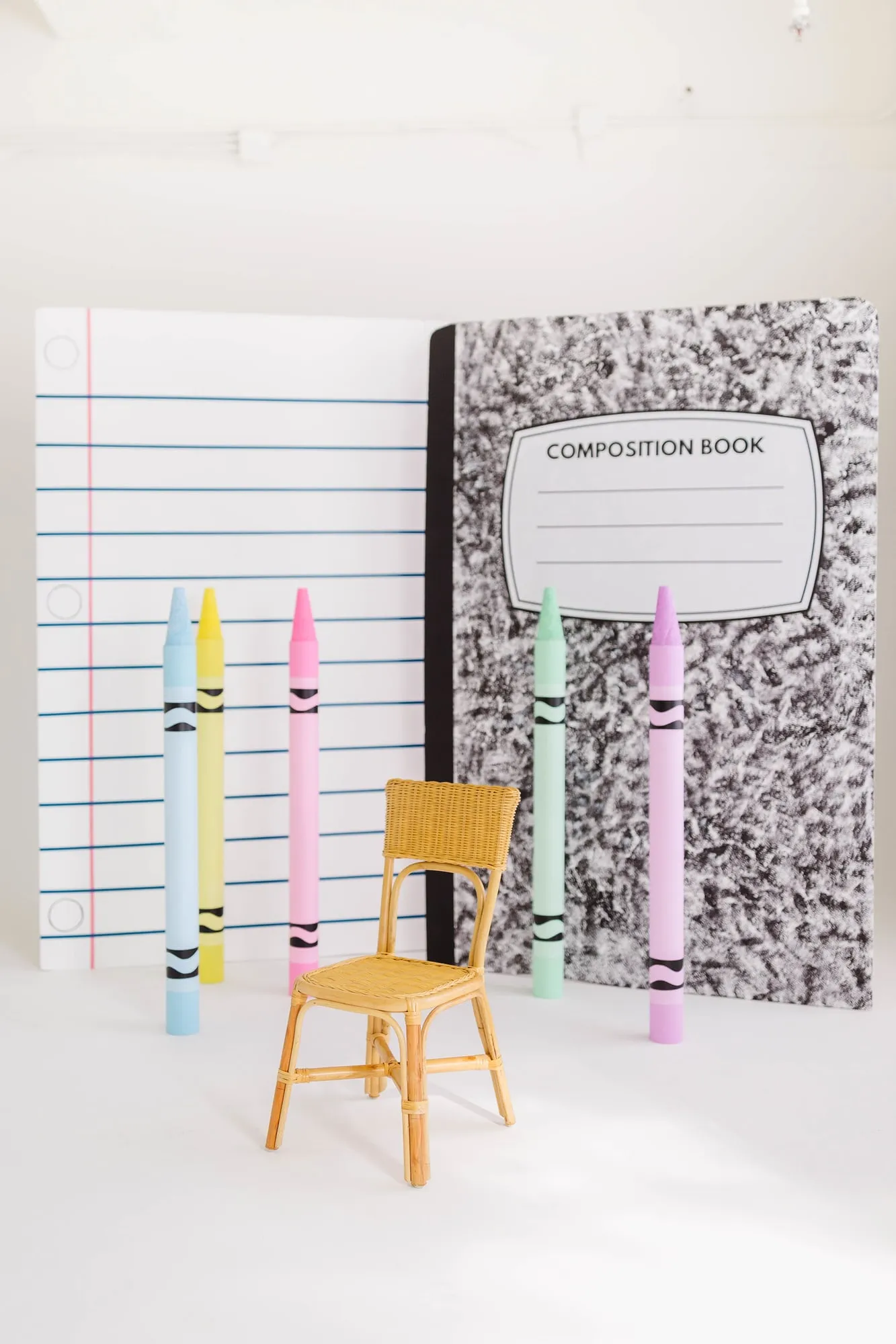 Back to school backdrop with notebook paper and composition book panels, giant pastel crayons and a rattan chair