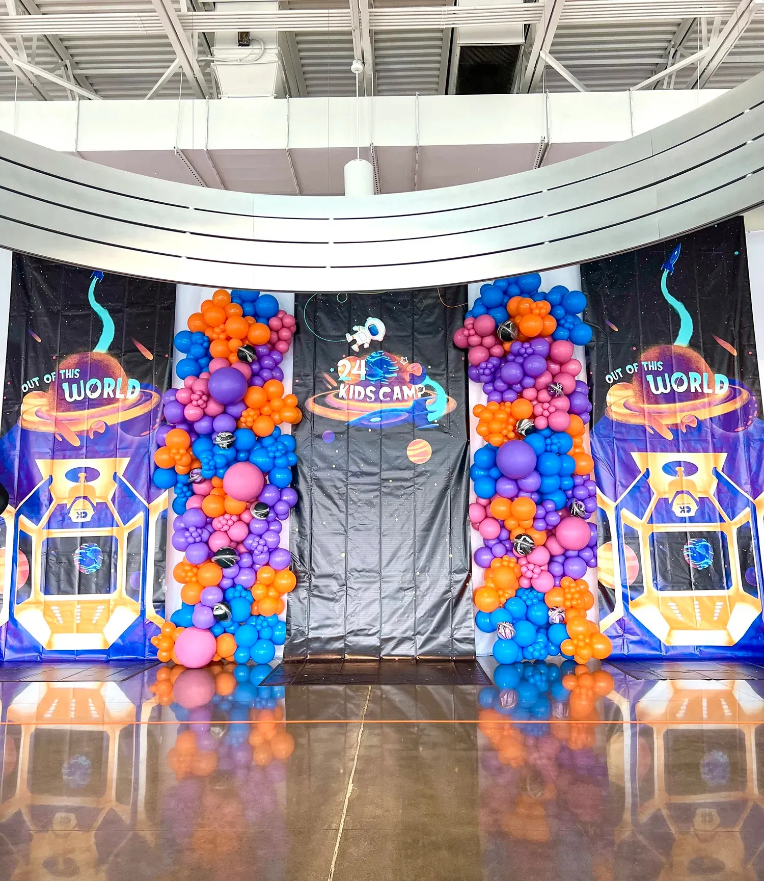 Space themed balloon columns in purple, orange, pink and blue beside a kids camp stage backdrop
