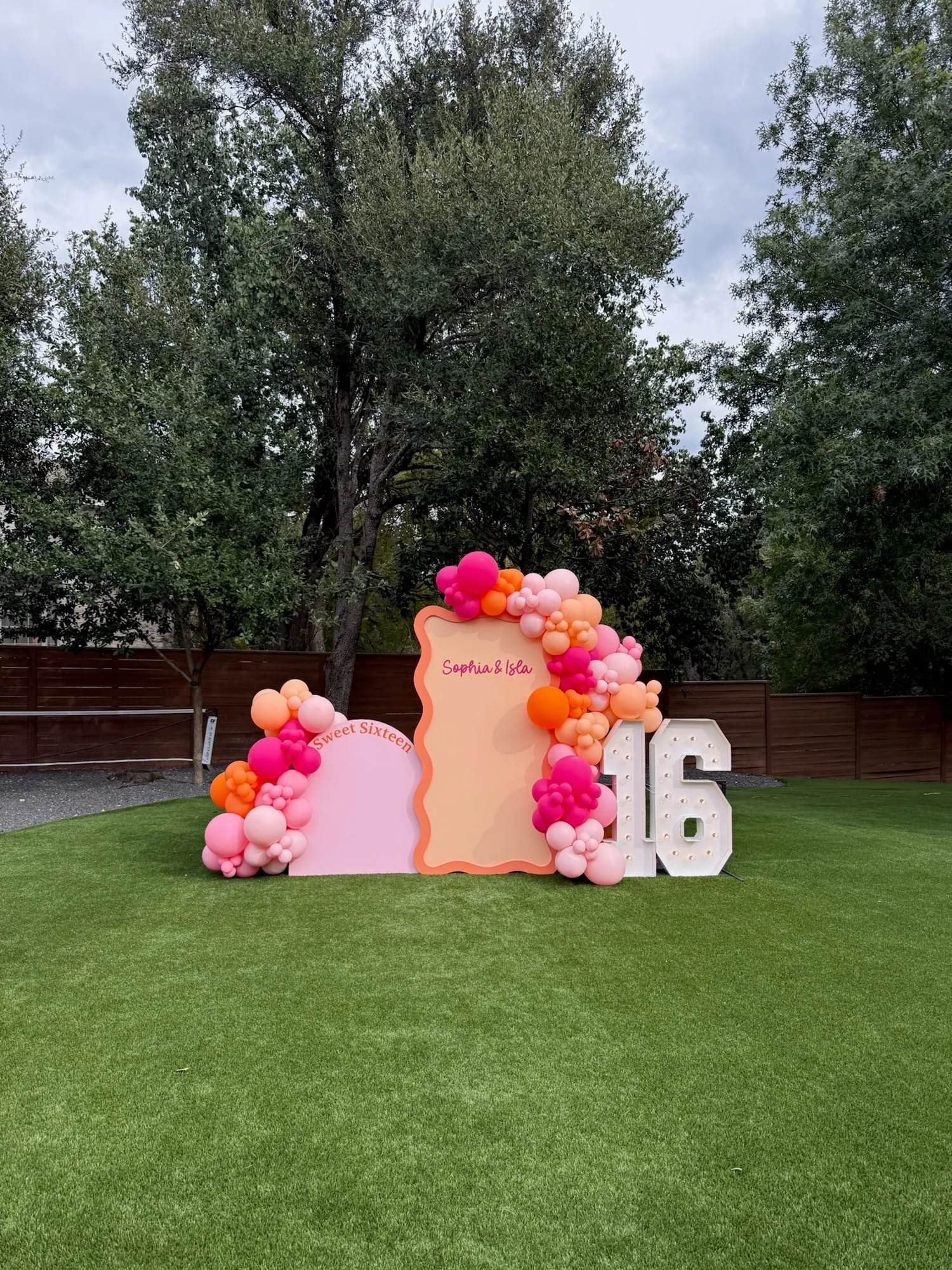 Sweet sixteen backdrop with pink and peach wavy arches and a hot pink, orange and blush balloon garland on the lawn