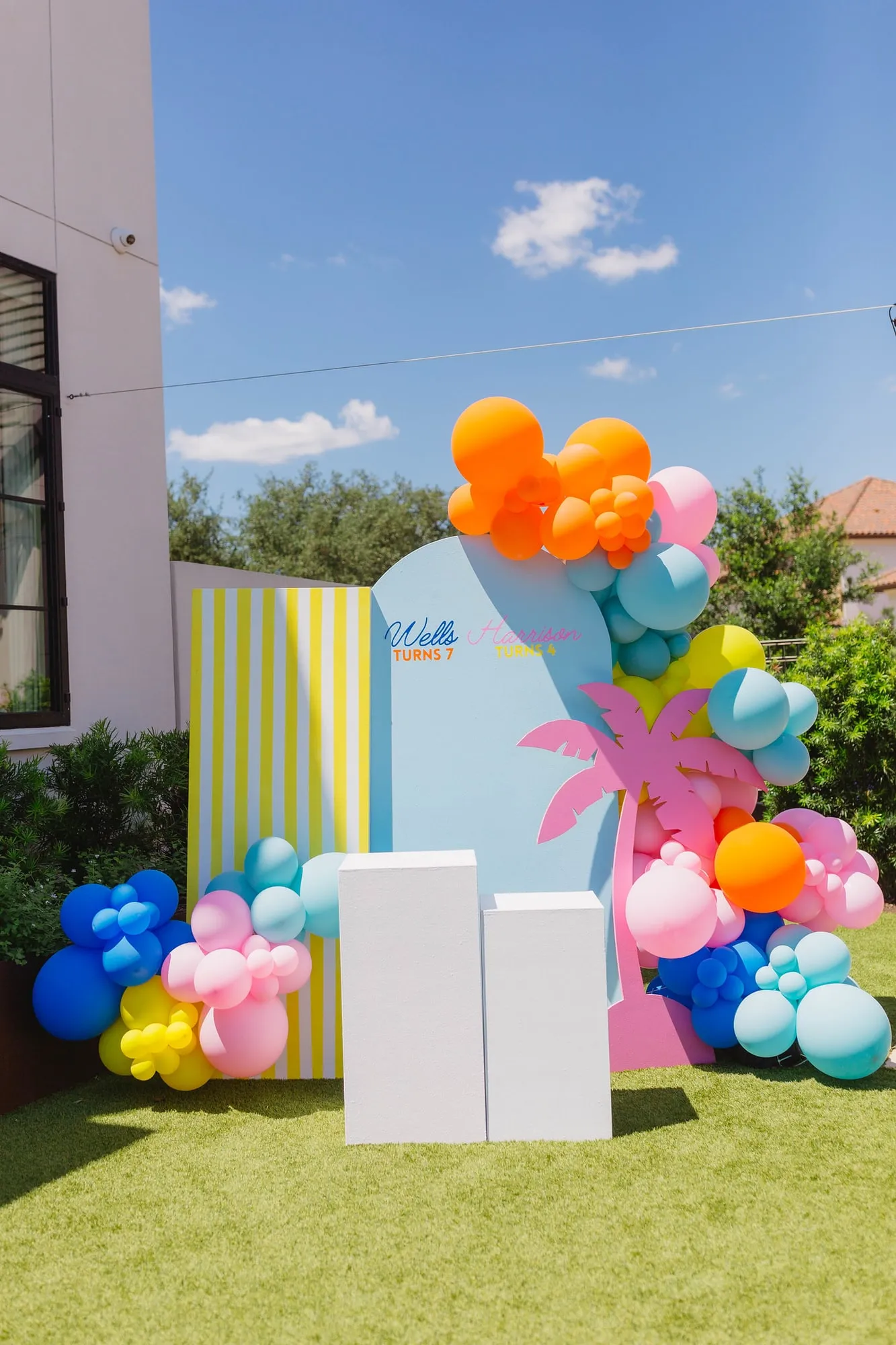 Tropical joint birthday with a blue arch, yellow striped panel, pink palm tree cutout, white pedestals and bright balloon garland